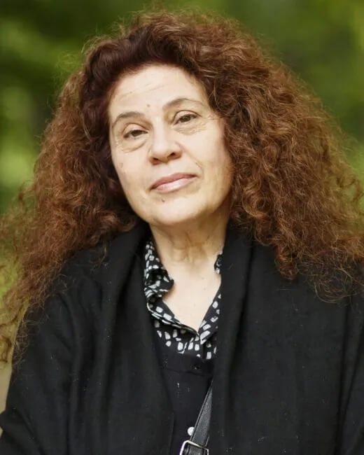 A woman with long, curly brown hair wearing a black coat and a black patterned shirt stands outdoors, looking at the camera with a gentle expression. The background is blurred greenery.