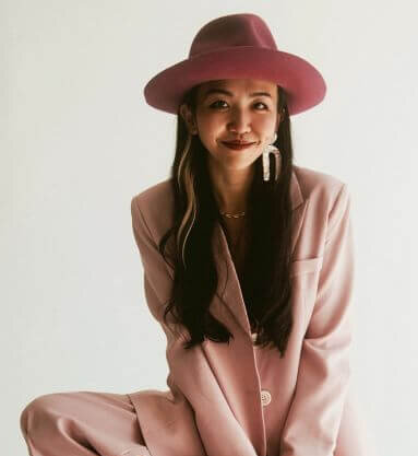 A woman wearing a wide-brimmed pink hat and a matching pink suit poses against a plain white background, smiling slightly.
