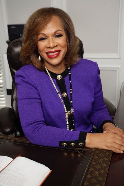 A woman with shoulder-length brown hair and red lipstick smiles while sitting at a desk. She is wearing a purple blazer with black trim and gold buttons, a pearl necklace, and has an open book in front of her.