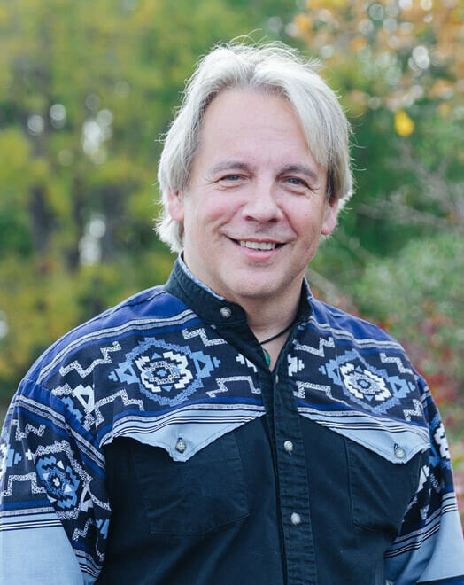 A smiling man with blonde hair stands outdoors, wearing a navy blue and black patterned button-up shirt with silver buttons. Green trees and foliage are blurred in the background.