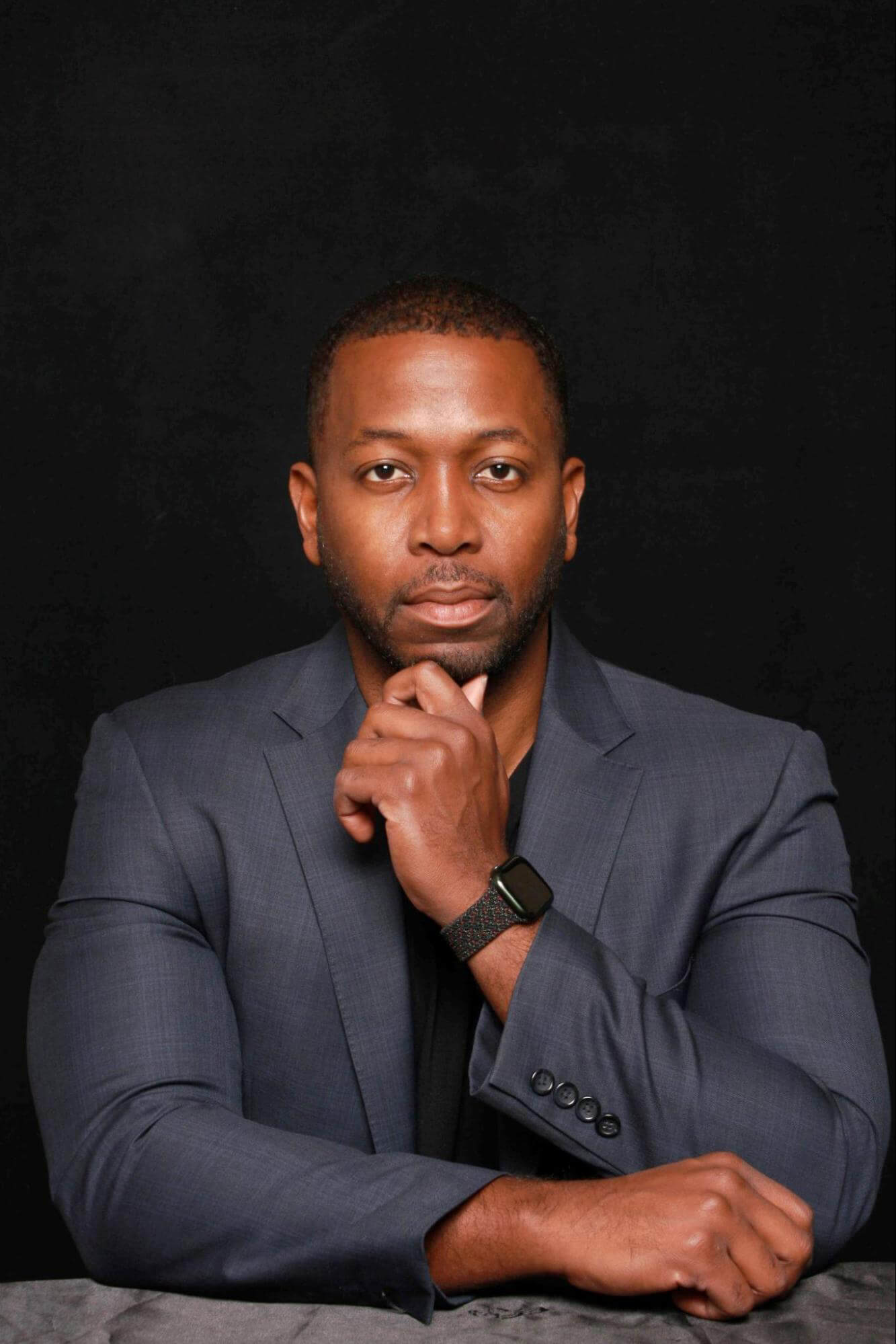 A man in a dark gray suit sits against a black background, resting his chin on his hand in a thoughtful pose. He wears a black shirt and a smart watch on his left wrist.