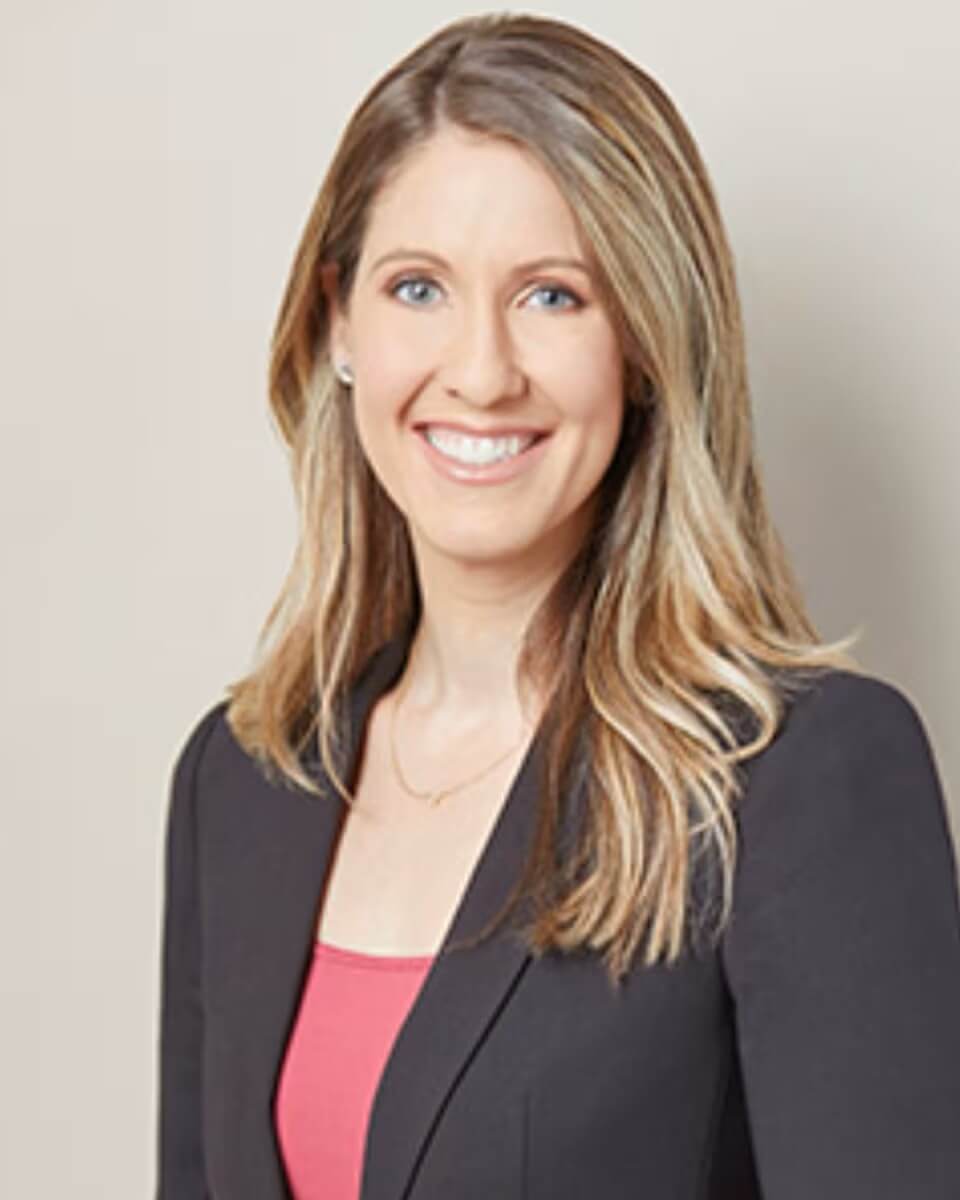 A woman with long, straight, light brown hair smiles at the camera. She is wearing a black blazer over a pink top and a delicate necklace, standing against a plain light background.
