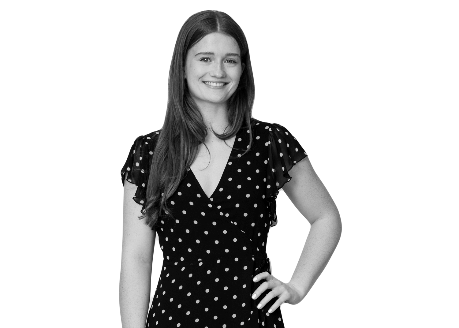 A young woman with long hair, wearing a black dress with white polka dots, stands smiling with one hand on her hip against a plain white background.