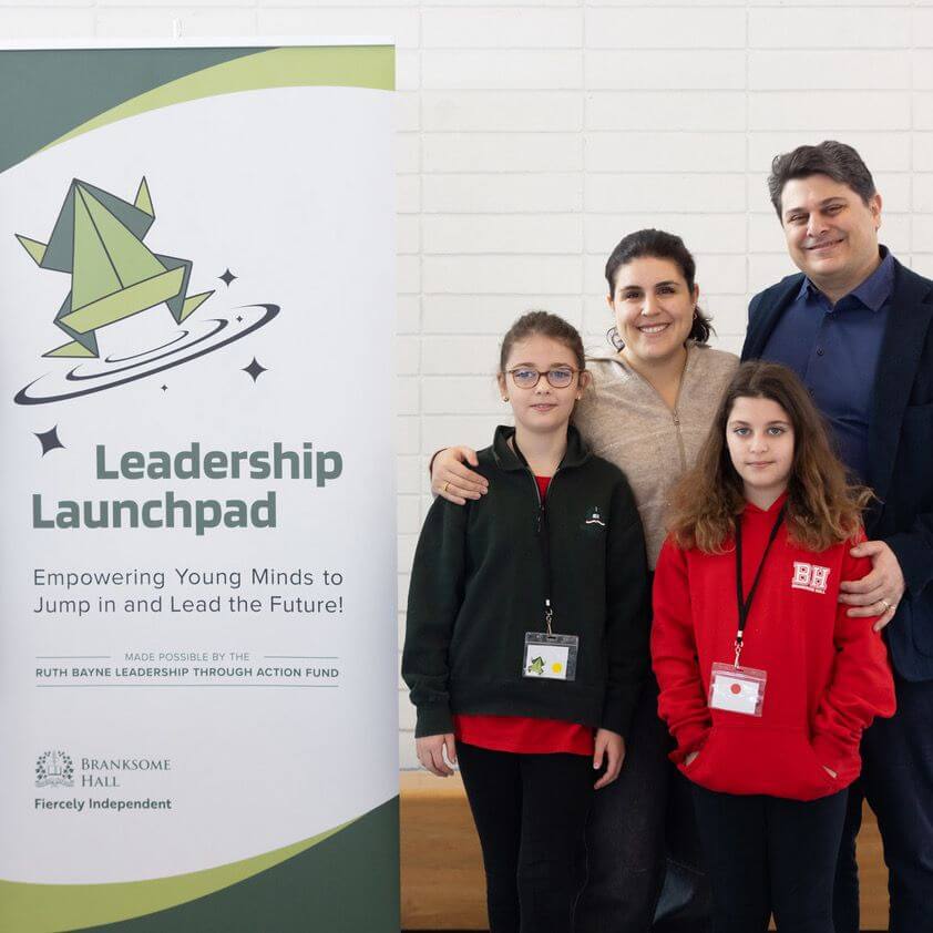 A family of four stands smiling next to a "Leadership Launchpad" banner, which features a green rocket illustration and promotes empowering young minds to lead the future. Two children wear school lanyards and sweatshirts.