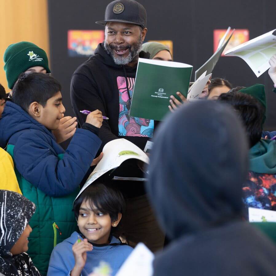 A smiling man in a cap holds books while surrounded by excited children, some holding open notebooks and pens, at what appears to be an indoor event or workshop.