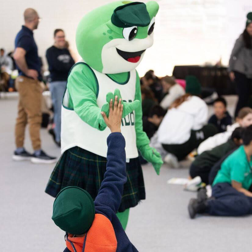 A person in a green frog mascot costume high-fives a child wearing a green hat and orange sweater, with a group of people seated on the floor in the background.