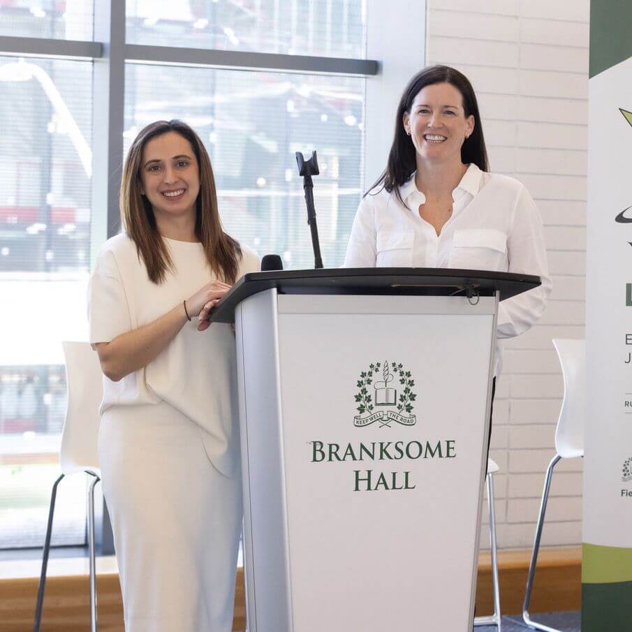 Two women stand smiling at a podium with a “Branksome Hall” sign. Both wear white outfits, and the background features large windows and a partial banner on the right.