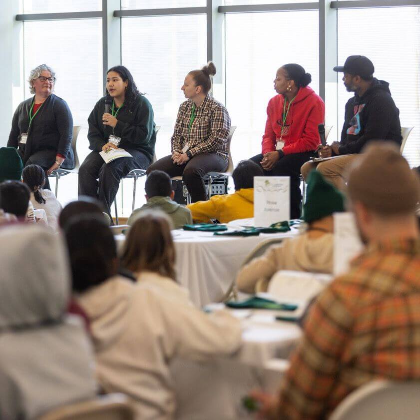 A panel of five people sits at the front of a room, speaking to an audience seated at round tables covered in white cloths. One panelist holds a microphone. Large windows let in natural light behind them.