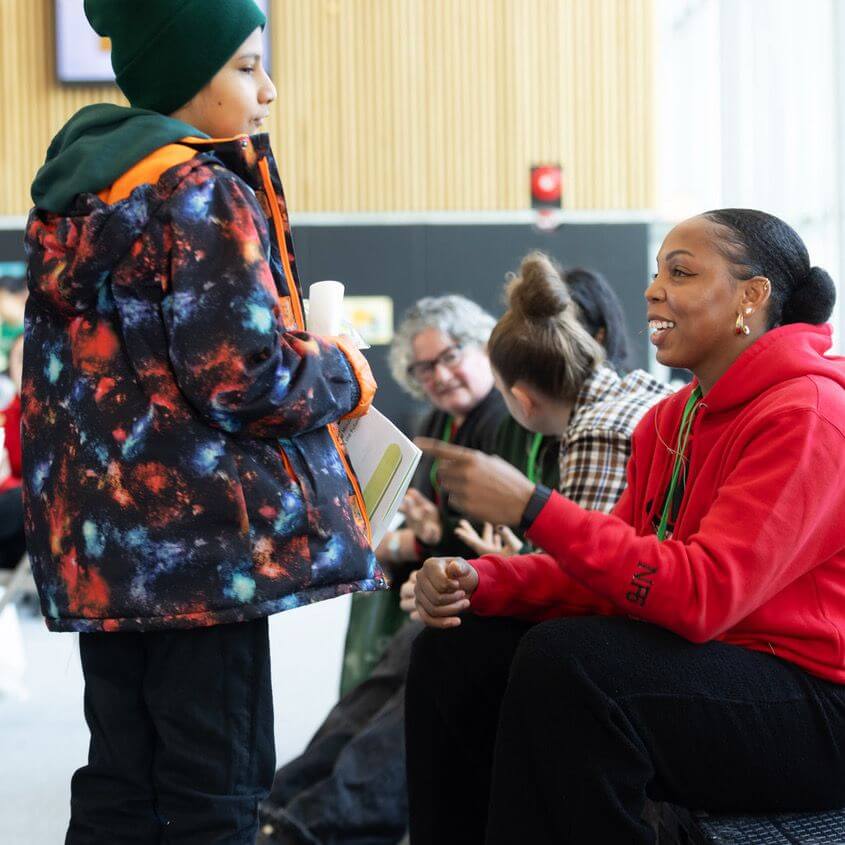 A child in a colorful jacket talks to a smiling woman in a red hoodie who is sitting down. Other people are sitting and talking in the background in a bright indoor space.
