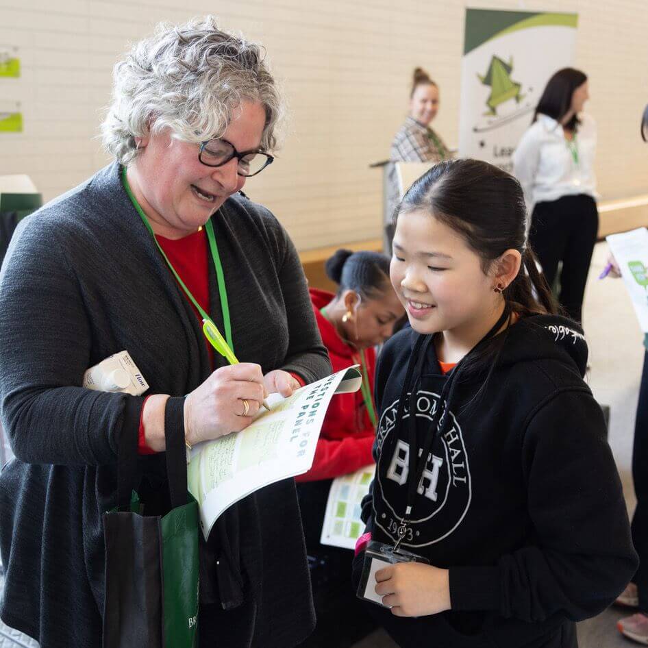 A smiling woman with gray curly hair signs a paper for a young girl in a black hoodie. Other people stand and sit in the background near a white brick wall at an indoor event.