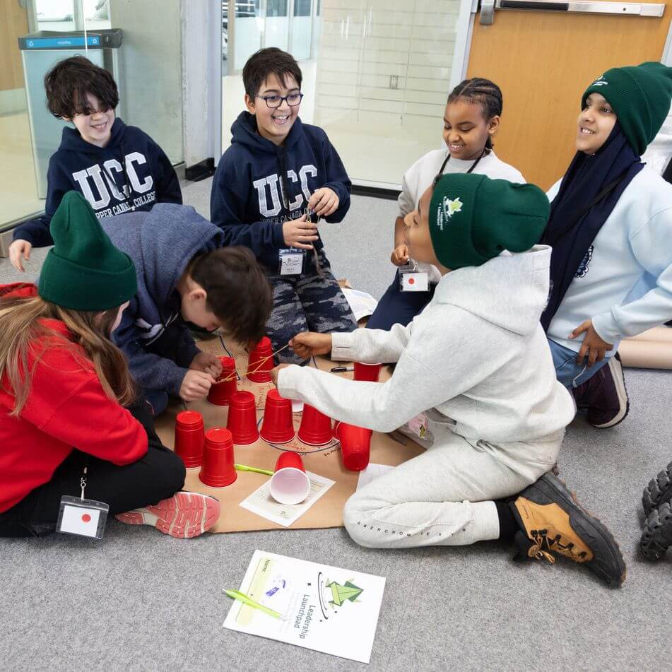 Seven smiling children sit in a circle on the floor, stacking red plastic cups as part of a group activity. They wear casual clothes, lanyards, and some have green beanies. A booklet lies on the floor nearby.