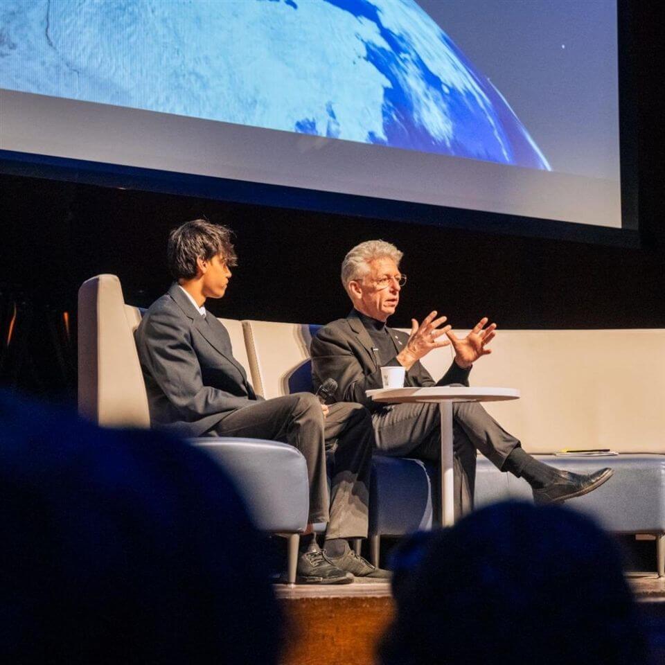 Two men in suits sit on a stage, engaged in conversation. One gestures with his hands while the other listens. Behind them is a large screen displaying an image of Earth from space.