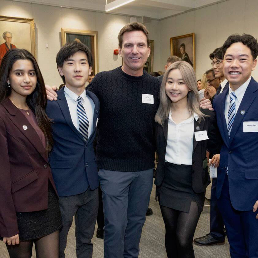 A group of five people, four young adults in business attire and one man in a sweater, pose together and smile at an indoor event with others and portraits in the background.