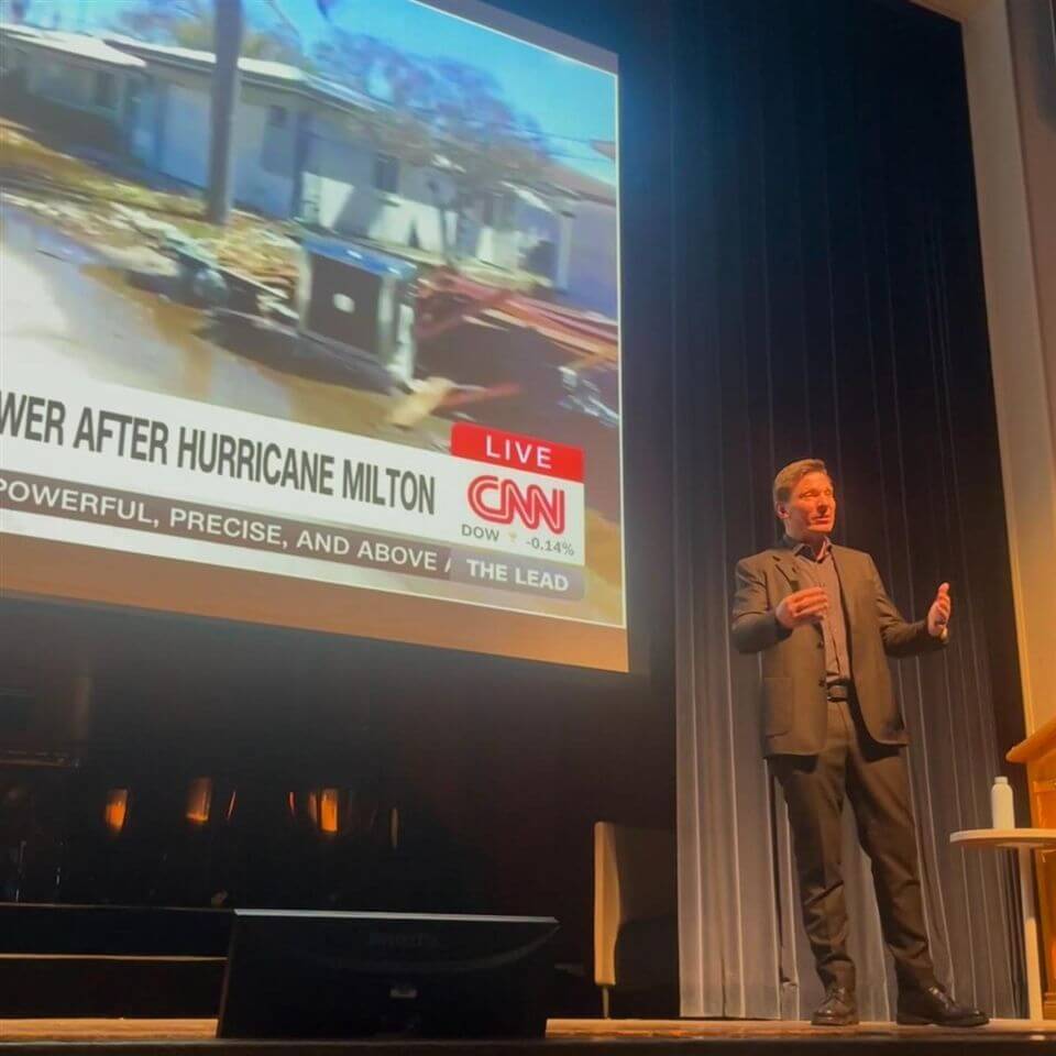 A man in a suit stands on stage giving a presentation, with a large screen behind him showing a CNN news broadcast about hurricane damage with the headline “Power After Hurricane Milton.”.