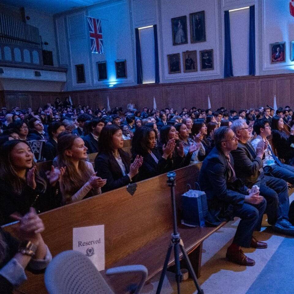 A diverse audience sits in wooden pews, attentively watching an event and clapping in a large hall adorned with flags and portraits on the walls. A "Reserved" sign is visible on one of the front seats.