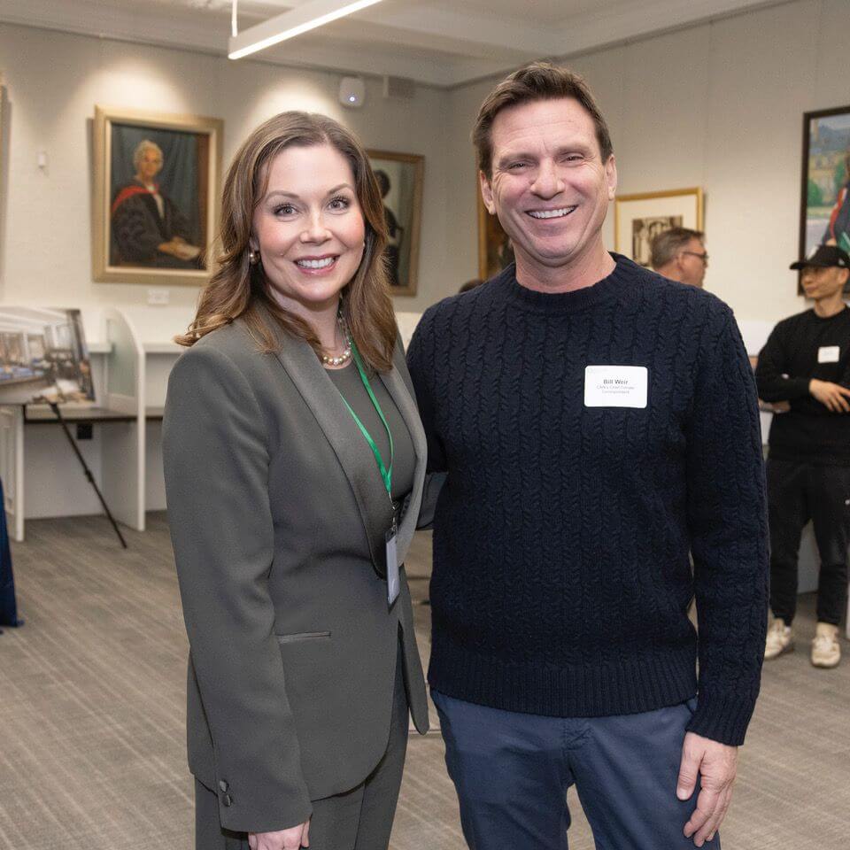 A woman in a green suit and a man in a dark sweater, both smiling, stand together in an art gallery with framed paintings on the walls and people conversing in the background.