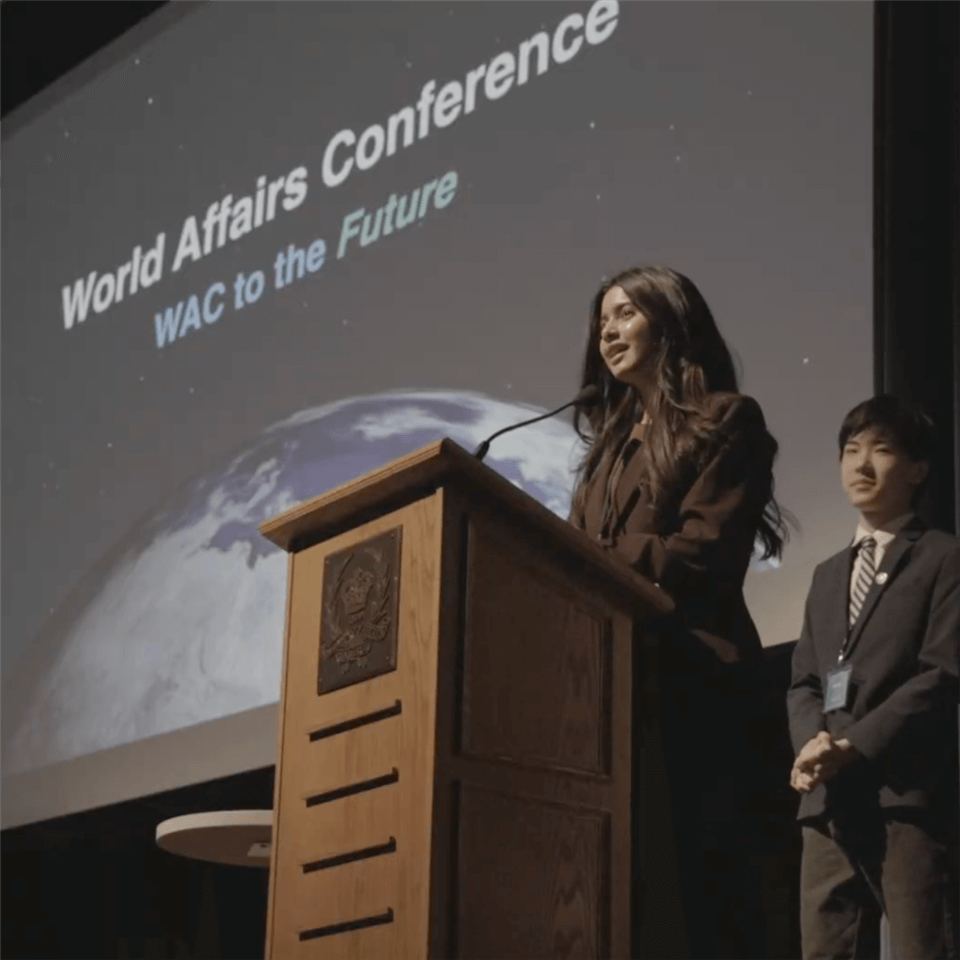 A woman speaks at a podium on stage, while a young man stands nearby. The screen behind them reads "World Affairs Conference: WAC to the Future," with an image of Earth in the background.