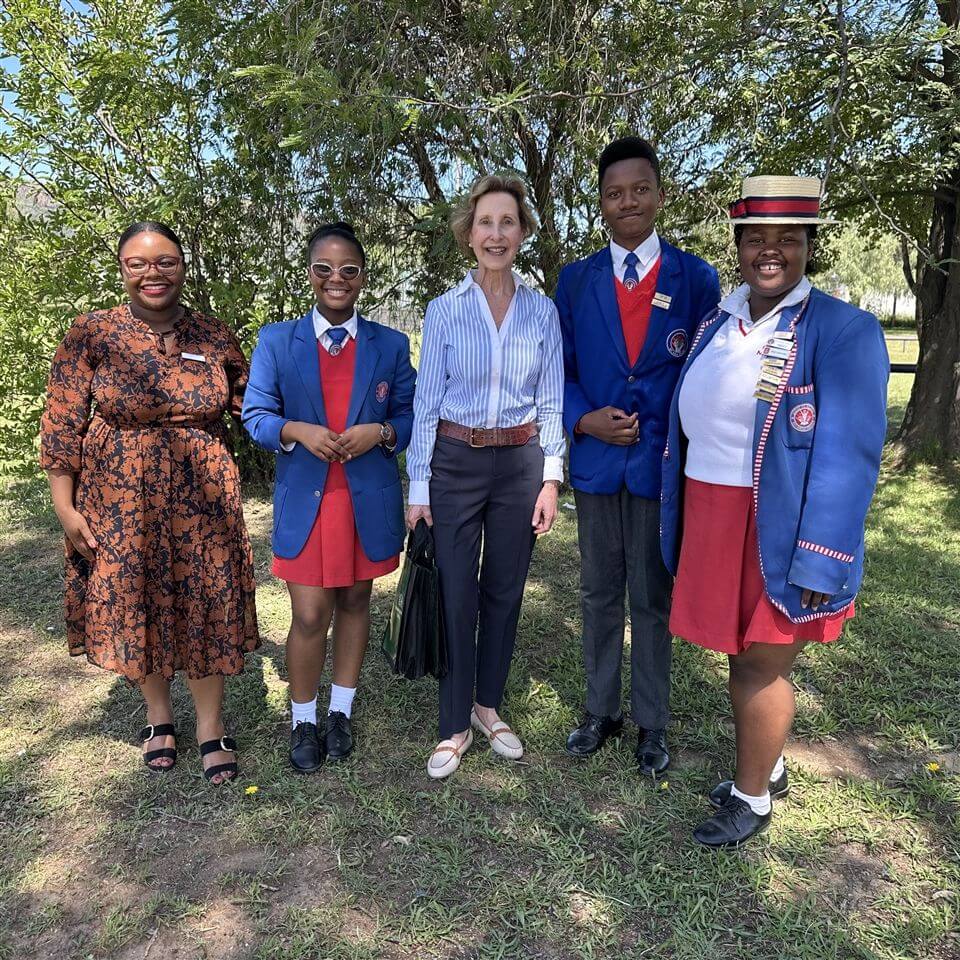 Five people stand outside on grass in daylight; four wear school uniforms with red skirts and blue blazers, one wears a floral dress. They are smiling, with trees and greenery in the background.
