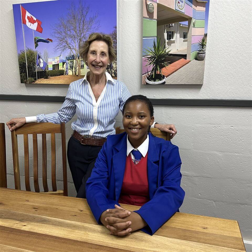 Two women pose in an office setting. One, standing, wears a light blue striped shirt; the other, seated, wears a blue blazer and red sweater. Framed photos of colorful buildings hang on the wall behind them.