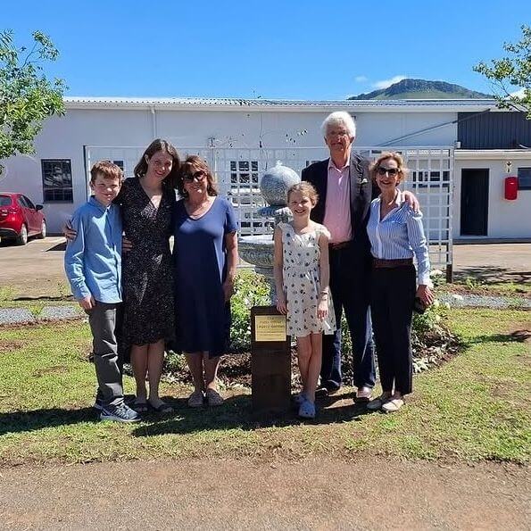 Six people, including two children and four adults, stand smiling together in front of a small garden with a plaque and a spherical sculpture, with a building and mountain in the background on a sunny day.