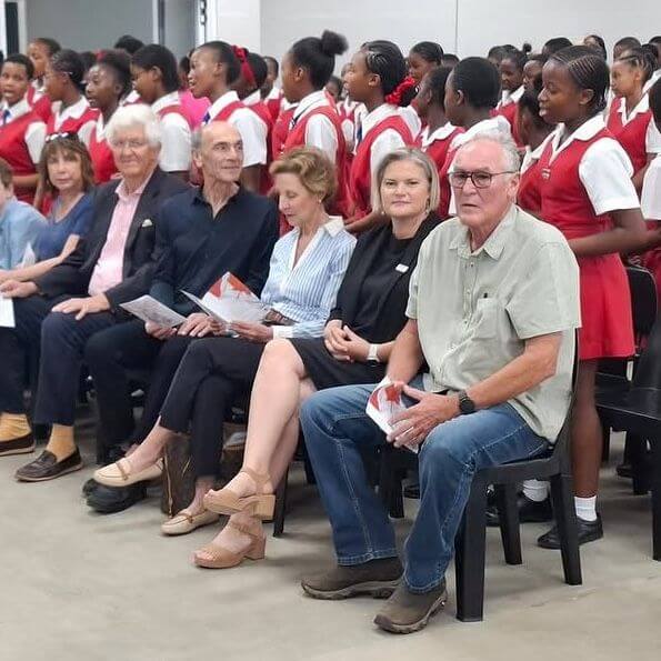 A group of adults sits in the front row of chairs, holding programs, while a choir of young girls in matching red and white uniforms sings behind them inside a large room.