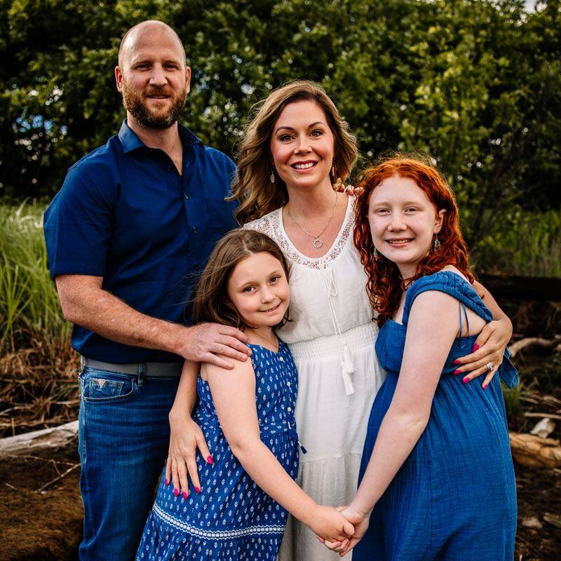 A family of four poses outdoors; a man and woman stand behind two young girls. The woman and girls have light brown or red hair and wear blue or white dresses, while the man wears a navy shirt and jeans. Green trees are in the background.
