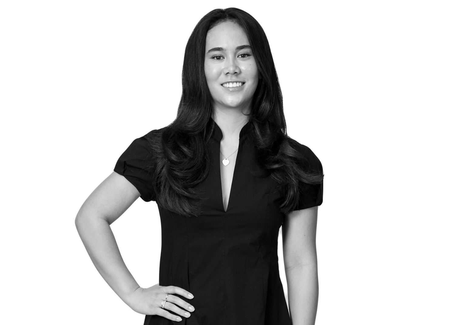 A woman with long dark hair, wearing a short-sleeved black dress, stands smiling with one hand on her hip against a plain white background.