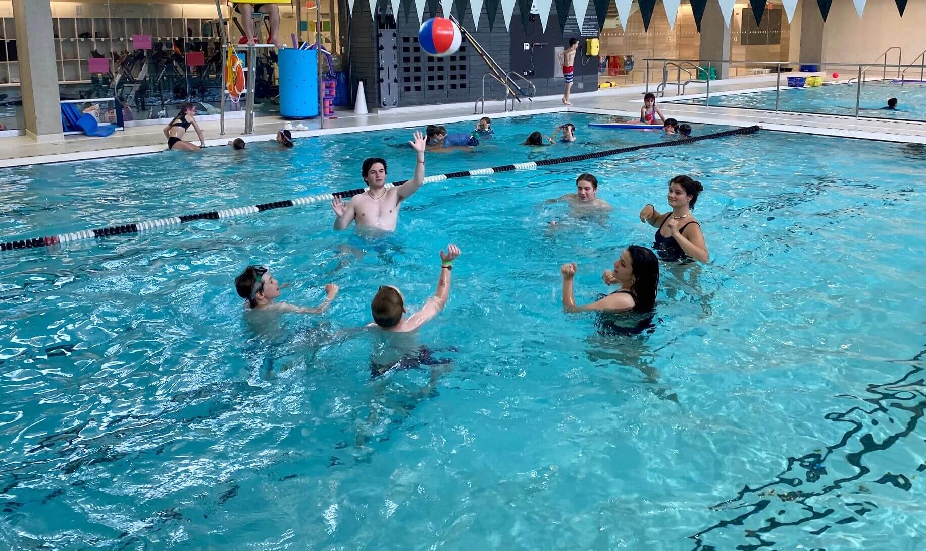 A group of people are playing with a beach ball in an indoor swimming pool. Some are splashing or reaching for the ball while others swim nearby. People are in swimwear and the pool area is well-lit.