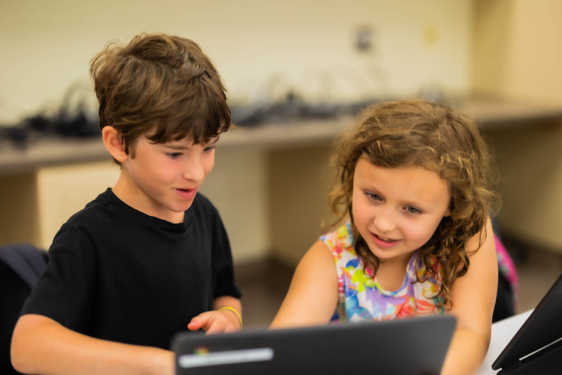Two young children, a boy in a black shirt and a girl in a colorful dress, are smiling and working together on a laptop in a classroom setting.