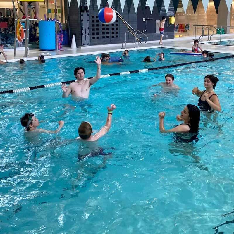 A group of people in a swimming pool play a game with a colorful beach ball. They are smiling and reaching for the ball, with more swimmers and pool equipment visible in the background.