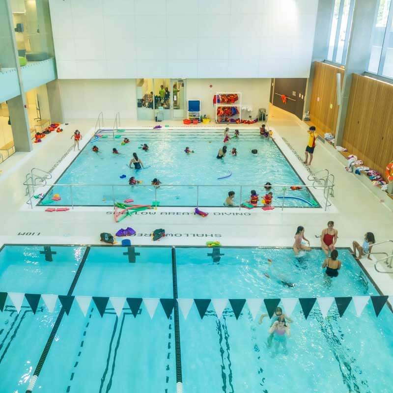 A bright indoor swimming pool with people of various ages swimming, playing, and standing around two connected pools; some are wearing swim gear, and an instructor stands near the poolside.