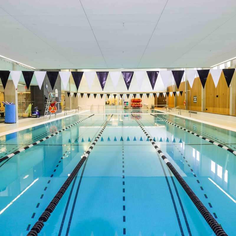 Indoor swimming pool with clear blue water, black lane dividers, and triangular black-and-white flags overhead. The pool area has wooden walls, bright lighting, and pool equipment visible in the background.