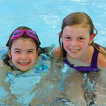Two young girls smiling while swimming in a pool. One girl wears pink goggles on her head, and the other has wet hair and a purple swimsuit. Both appear happy and are enjoying the water.