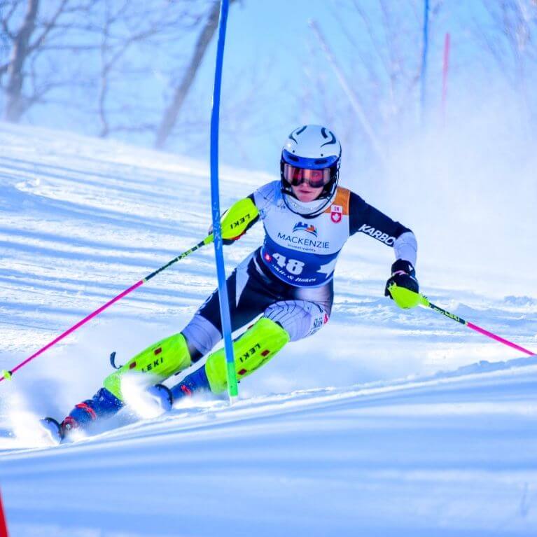 A skier in full gear, including a helmet and goggles, races downhill around a slalom gate on a snowy slope, kicking up powder under clear blue skies.