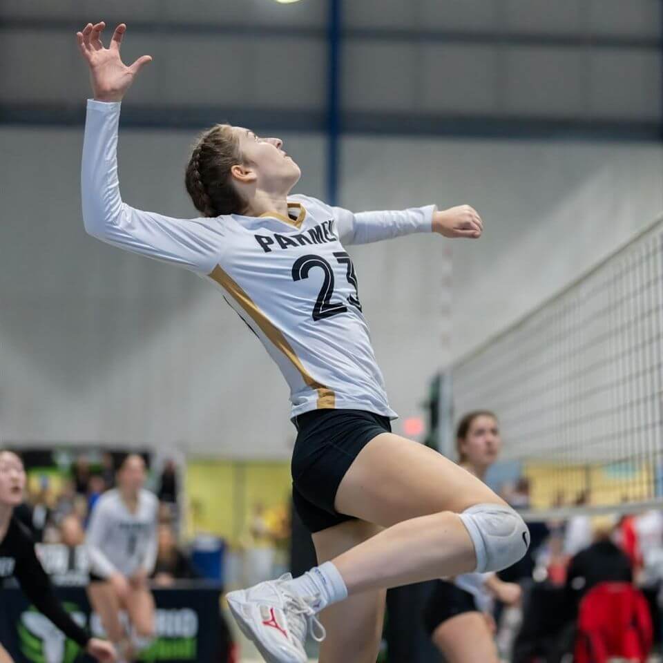 A volleyball player in a white and black uniform with the number 23 prepares to spike the ball during an indoor match. She is mid-air near the net, with her teammates and opponents visible in the background.