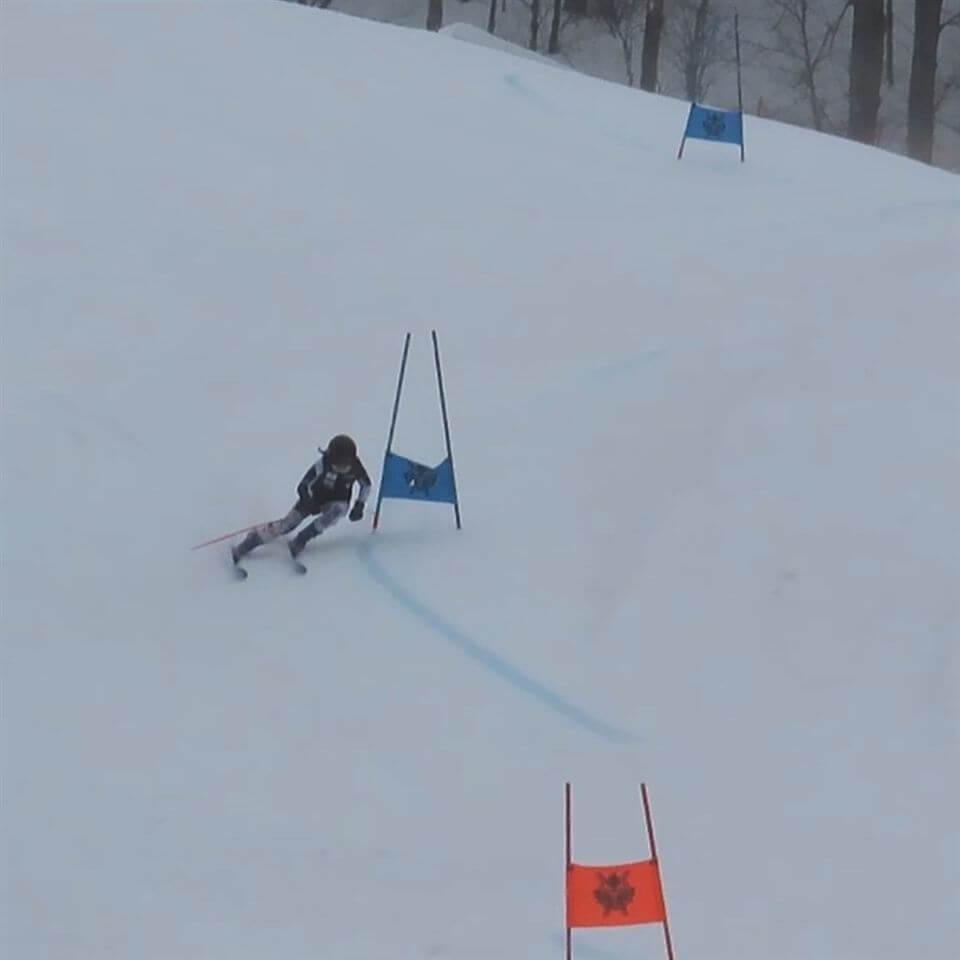 A skier in black and white gear navigates a slalom course on a snowy hill, turning past a blue gate, with another blue gate ahead and an orange gate in the foreground. Trees are visible in the background.