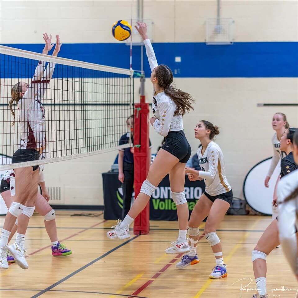 A volleyball player in a black-and-white uniform jumps to spike the ball over the net as an opponent tries to block; teammates and officials watch on in a gymnasium.