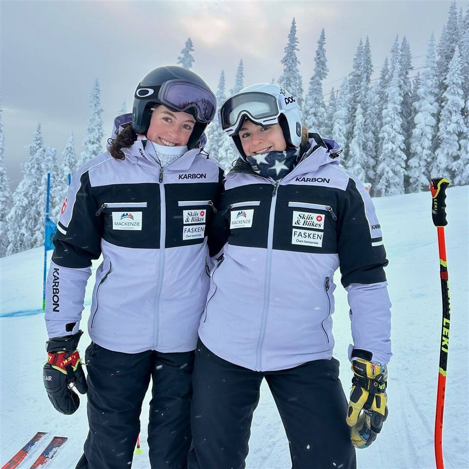 Two skiers in matching winter jackets and helmets stand close together, smiling on a snowy slope with ski poles, surrounded by snow-covered trees and skis lying on the ground.