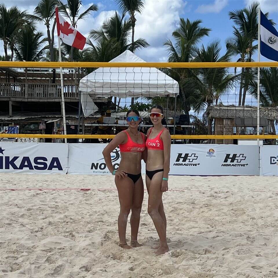 Two female beach volleyball players in matching red tops and black bottoms stand on a sandy court with a volleyball net behind them. Canadian and Salvadoran flags are visible in the background, along with palm trees and a blue sky.