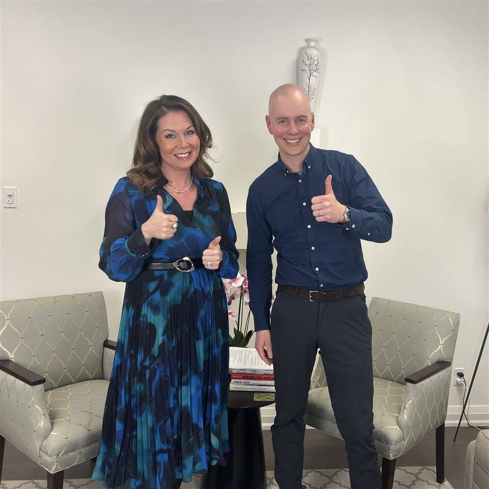 Two people stand smiling and giving thumbs up in a bright room with two patterned chairs, a small table with books, and a plant in the background. The woman wears a blue dress; the man wears a dark shirt and pants.