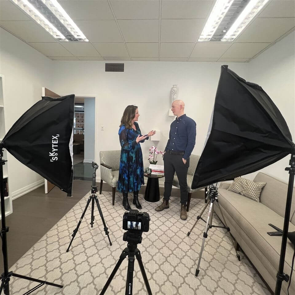 Two people stand and talk in a well-lit office, surrounded by photography lights and a camera on a tripod. They appear to be preparing for or filming an interview or video.