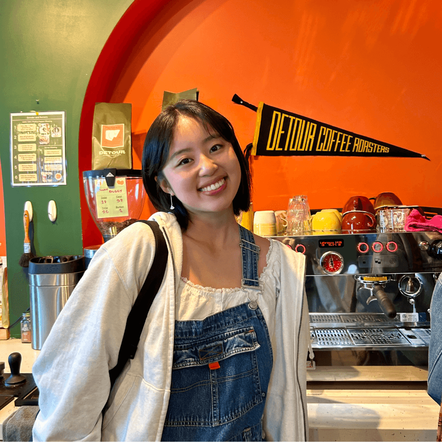 A smiling woman with short dark hair stands in a coffee shop wearing denim overalls and a white top. Behind her are coffee equipment, colorful mugs, and a yellow "DETOUR COFFEE ROASTERS" pennant on an orange wall.