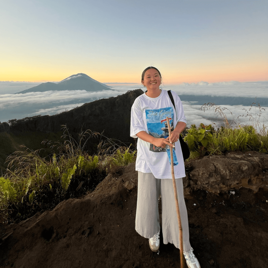A smiling person stands on a mountain trail at sunrise, holding a walking stick. Lush greenery and clouds surround them, with a distant volcano visible against a colorful sky.