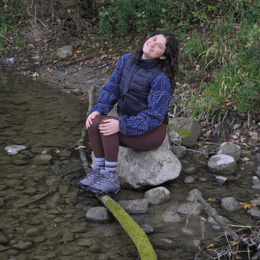 A woman with curly hair, wearing a blue patterned jacket, brown leggings, and hiking boots, sits on a large rock by a clear, shallow creek surrounded by greenery and stones, smiling and looking up.