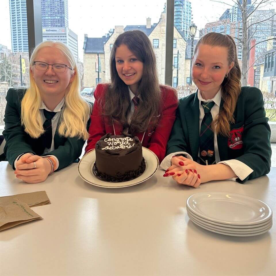 Three young women in school uniforms sit at a table. The girl in the center holds a chocolate cake with "Congrats Work December" written on it. They are smiling, with plates stacked on the table in front of them. City buildings are visible outside.