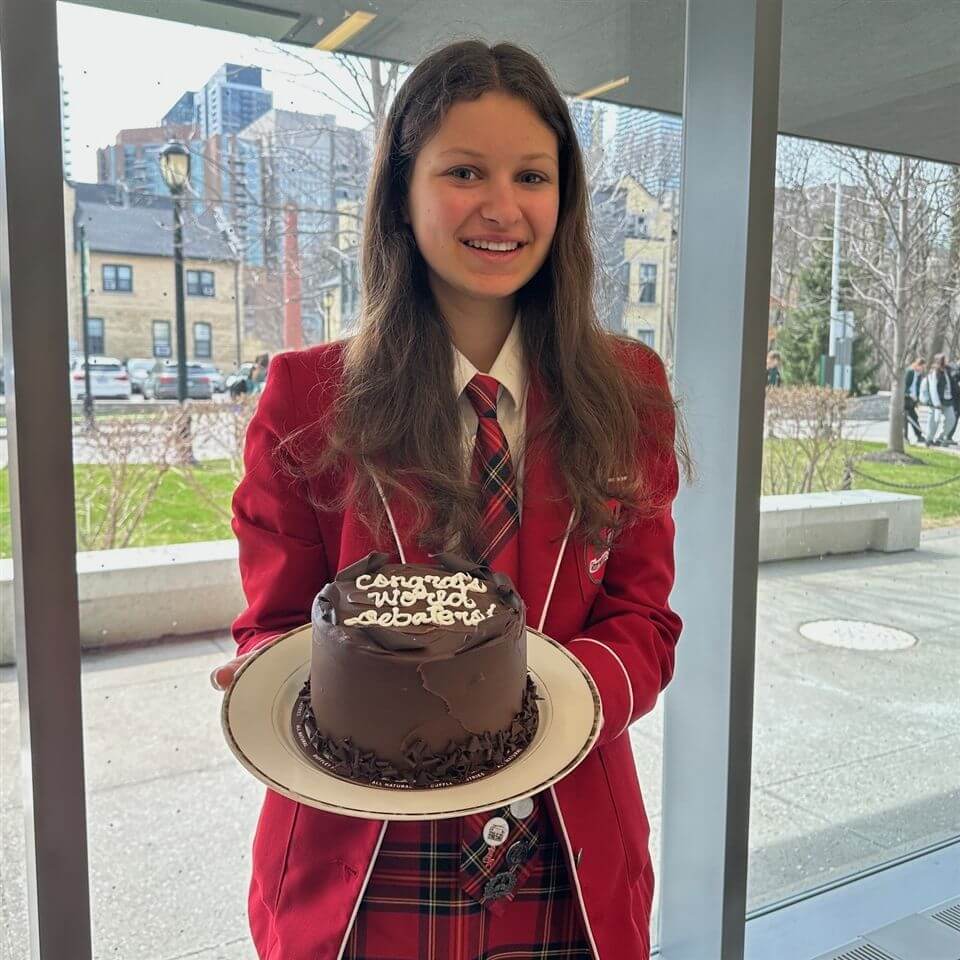 A smiling girl in a red school uniform holds a chocolate cake with “Congrats, you did it, Sabatier!” written on top. She stands indoors near large windows with a cityscape and trees visible outside.