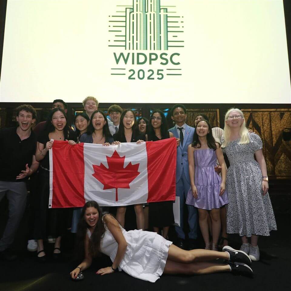 A group of smiling young people pose together, holding a Canadian flag. Behind them is a large screen displaying the logo and text “WIDPSC 2025.” One person lies in front of the group, smiling at the camera.