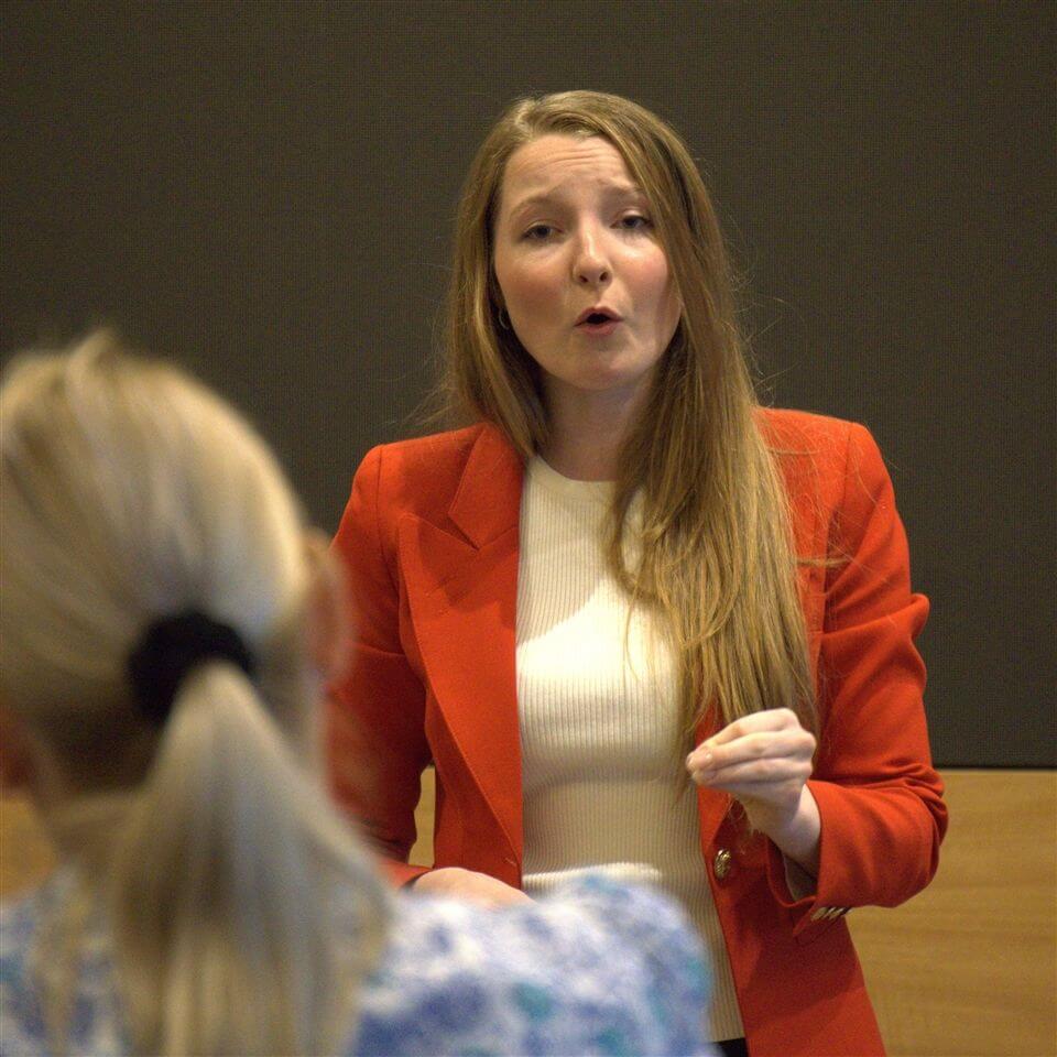 A woman in a red blazer and white top is speaking passionately, facing an audience. Another person, seen from behind, is listening attentively. The background is plain and dark.