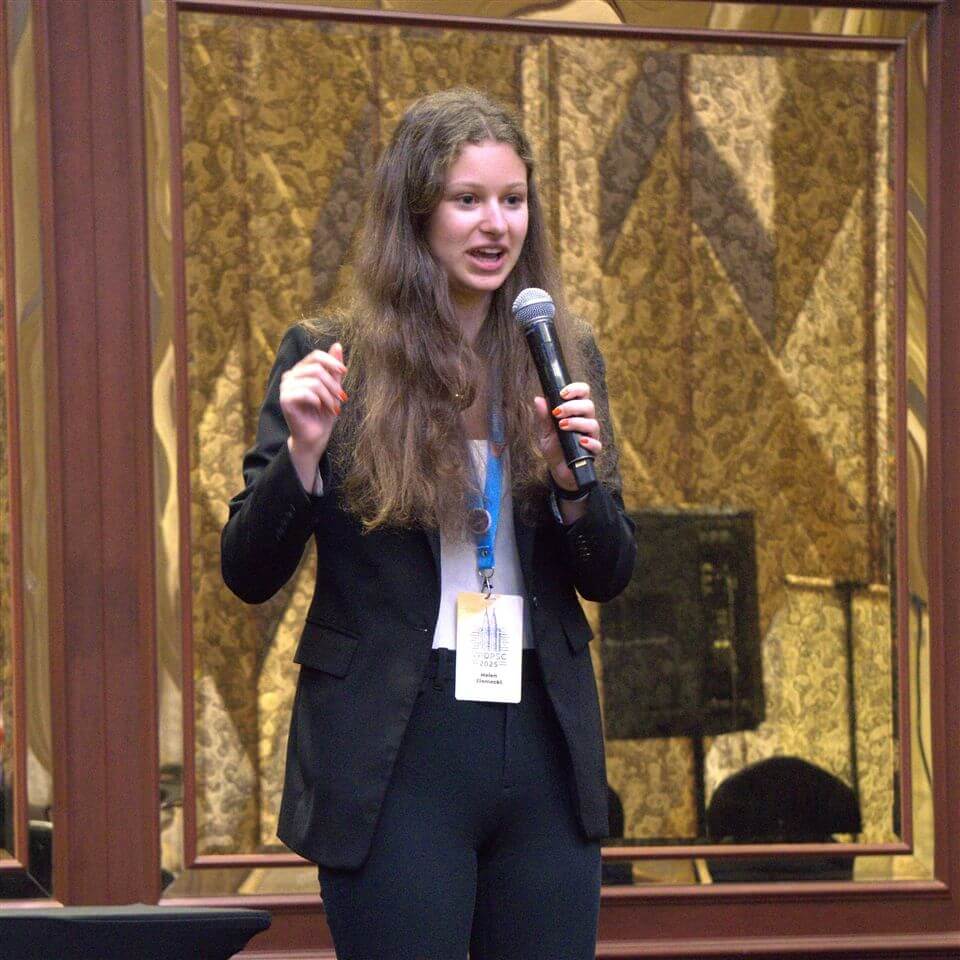 A woman with long brown hair, dressed in a black blazer and trousers, speaks into a microphone while gesturing with her hand. She wears a name badge and stands indoors in front of ornate patterned curtains.