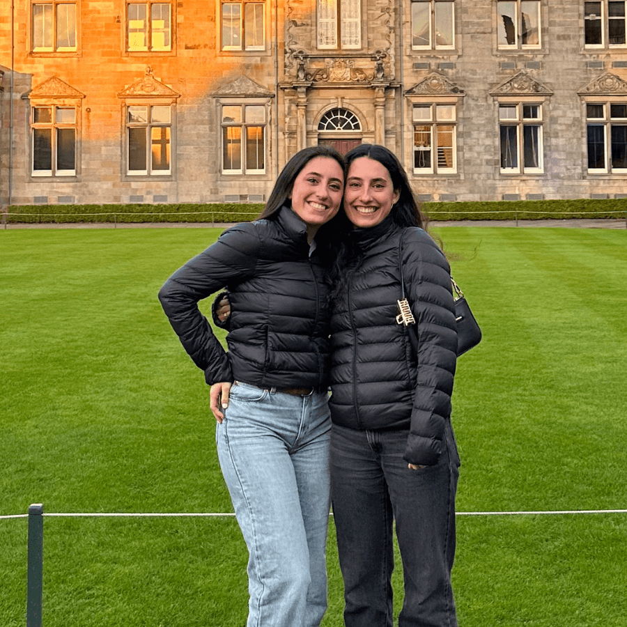 Two people wearing black puffer jackets and jeans stand smiling with their arms around each other on a green lawn in front of a large stone building with many windows.